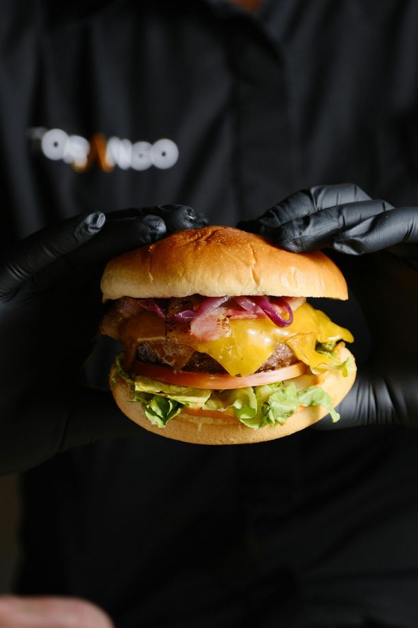 Hamburguesa sostenida por manos con guantes negros. Pan, queso, cebolla roja, tomate y lechuga visibles.